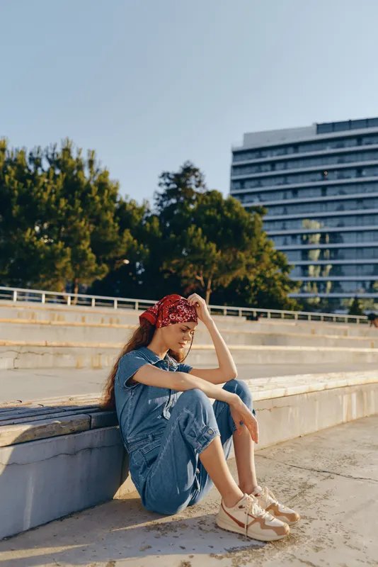Lifestyle woman in modern boho-western denim street style wearing a red bandana sits outdoors on concrete steps in warm sunlight with film color tones and casual sneakers.