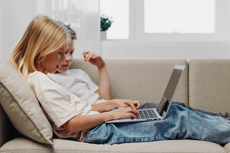 Two children sitting closely on a beige sofa using a laptop together in a bright room. Concept of learning, technology, childhood, and shared digital experience at home.
