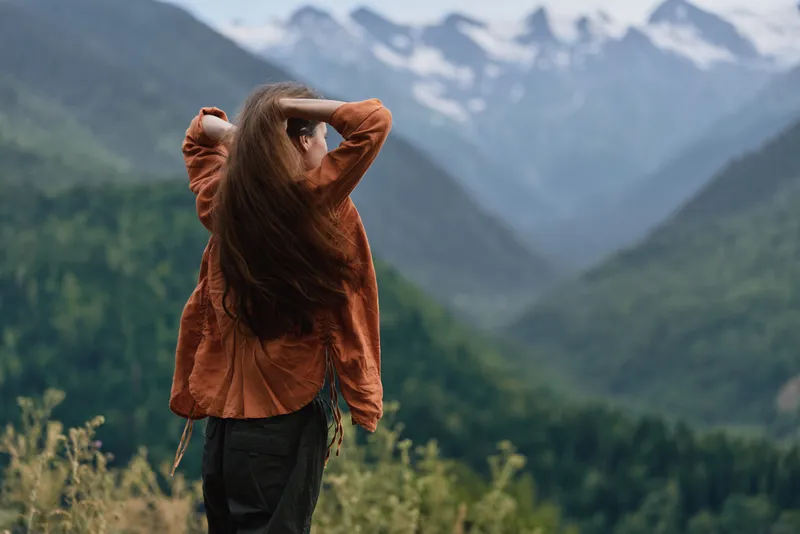 Woman stands outdoors with back to camera in a brown jacket, gazing at expansive mountains and lush valley scenery, creating a sense of freedom and peaceful adventure in nature.