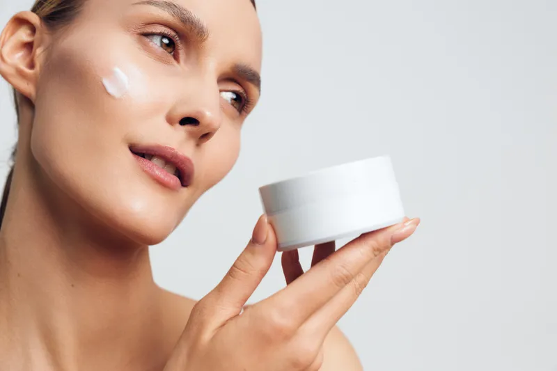 A young woman with clear skin applies white cream on her cheek holding a white jar close to her face. Concept for skincare, beauty routine, hydration, and self-care.