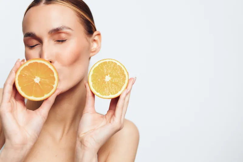 A young brunette woman closes her eyes and smiles softly as she holds two orange halves near her lips, creating a playful citrus moment against a clean white background.