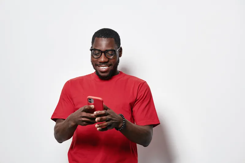 African American man with dark skin wearing glasses and red t shirt smiling while using smartphone for texting or social media on white background in casual style.