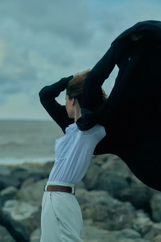 Model lifts flowing cape on windy beach, white shirt and light trousers, dramatic motion by the coast, ocean and rocks in the background, moody sky