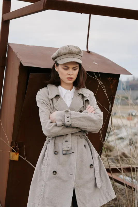 Young woman in beige trench coat and matching cap stands outside with arms crossed, looking serious. Rustic metal structure and dry plants in the background create moody atmosphere.
