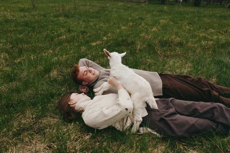 Goat kid interacts playfully with a couple lying on a grassy field, outdoor lifestyle moment, warm light, relaxed posture, soft breeze, natural colors, candid and tender scene