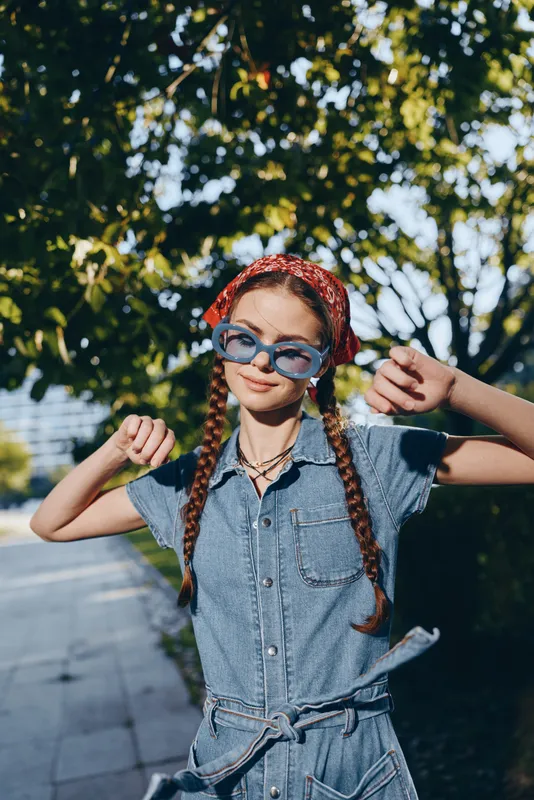 Lifestyle woman in modern boho-western denim street style smiles confidently wearing sunglasses and a red bandana with sunlight and lush greenery background.