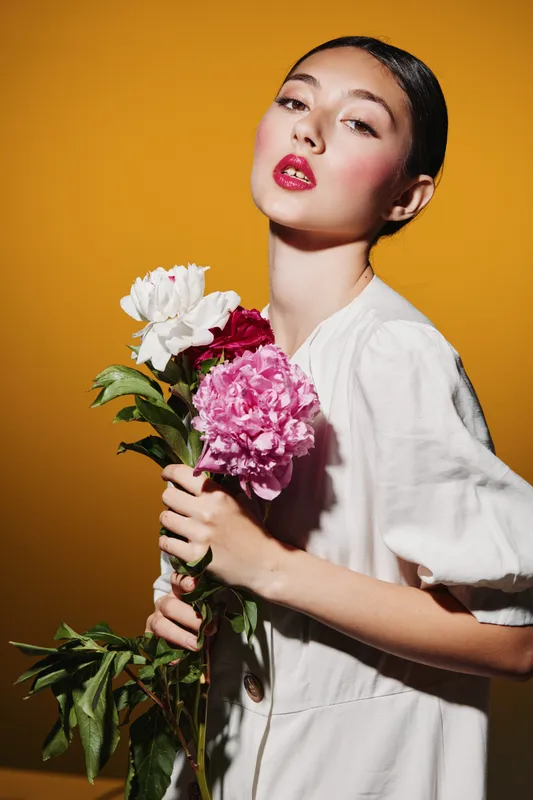 Young woman in a white dress holds pink peony and white roses against an orange background. Fashion creative beauty concept with elegant makeup and colorful flowers.