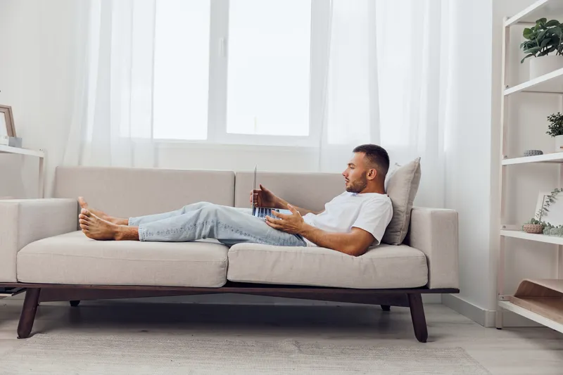 Relaxed man enjoying his free time on a comfortable sofa, dressed casually in a T-shirt and jeans, surrounded by a bright and airy living space.