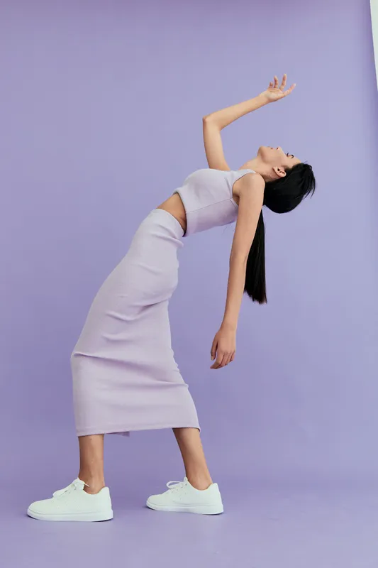 Young woman in lilac ribbed dress posing creatively against pastel purple background, showing graceful body movement and modern casual style with white sneakers.