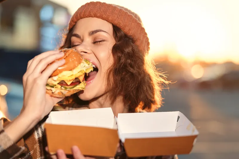 woman burger takeout urban smile joy candid authenticity. Young woman biting a large burger from a takeout box on a city street at golden hour glow, candid lifestyle, emotional storytelling.