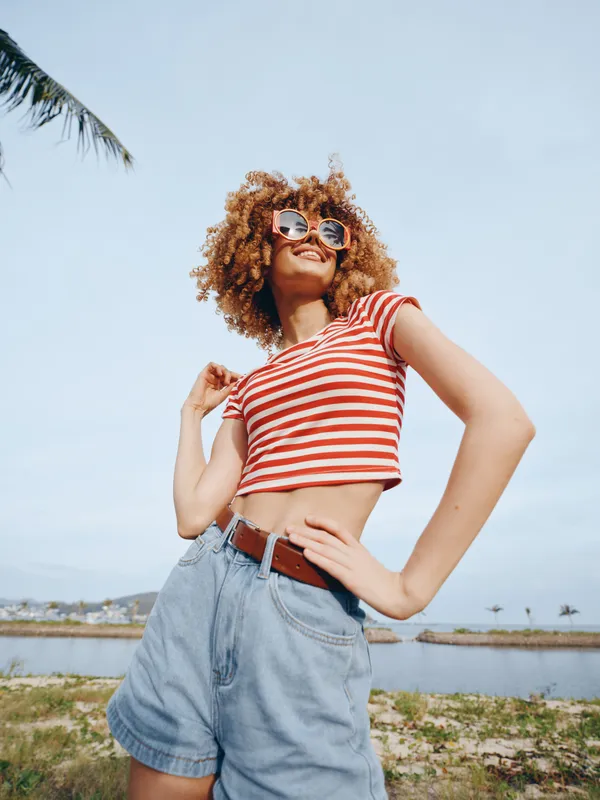 A young woman with curly hair wearing a red striped crop top and denim shorts stands outdoors under a bright sky, exuding a carefree summer vibe.