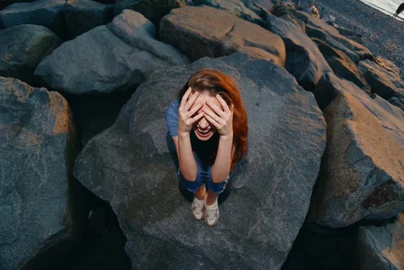 frustrated woman sitting on large rocks covering her face with hands in casual clothes outdoors by the water