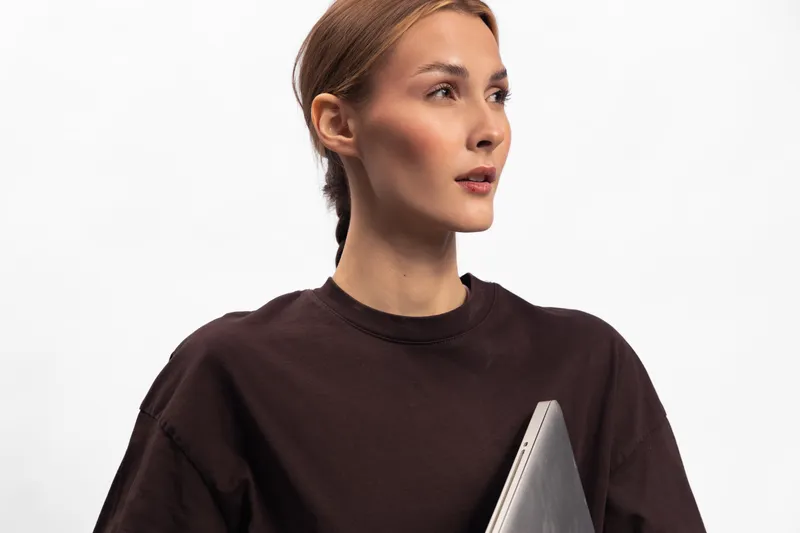Young woman in dark sweater holds laptop and looks aside on white background. Concept of technology, business, and modern lifestyle with confident expression and minimalistic style.