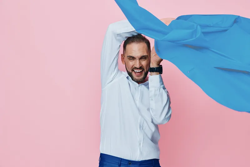 A professional man in a white shirt stands against a solid pink backdrop, raising his arms with a blue fabric flying through the air, exuding confidence, energy, and dynamic mood.