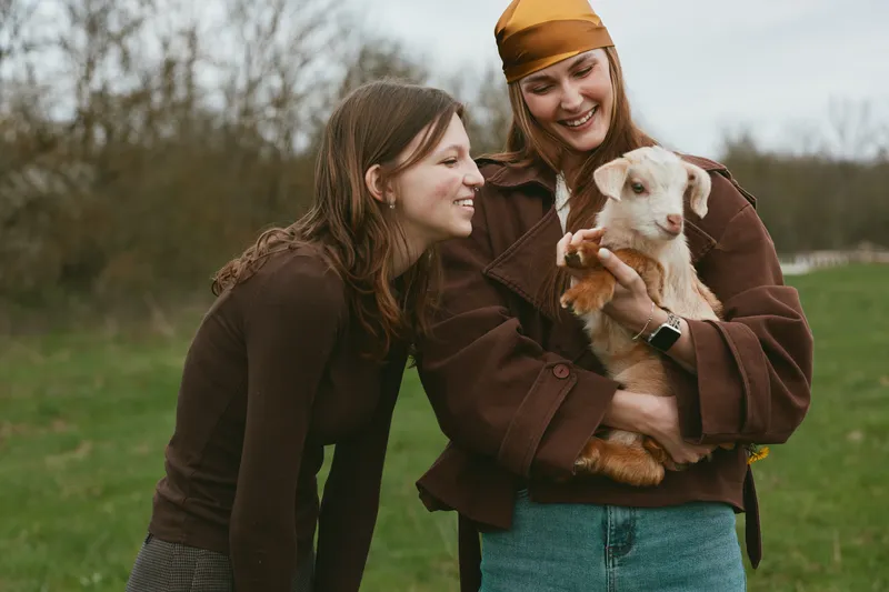 Two friends laugh outdoors as they cradle a goat kid, sharing smiles in a sunlit countryside scene, a wholesome lifestyle moment that highlights friendship, animal bonding, care, joy, and everyday