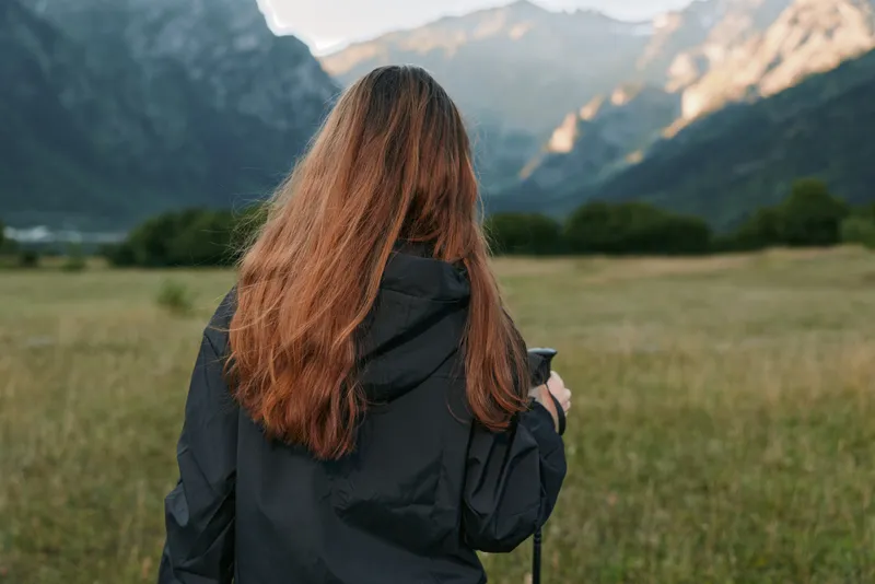 A person with long hair wearing a dark jacket stands in a windswept meadow, facing distant mountains. They hold a trekking pole as soft light bathes the landscape, creating a calm outdoors mood.