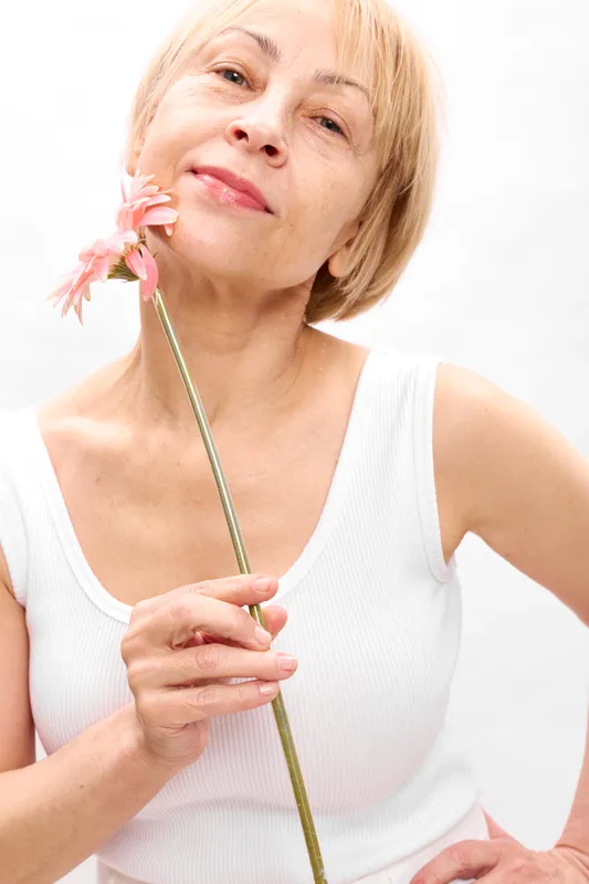 Senior blonde woman in studio, white background, with a smile, holding a flower stem near lips as if tasting, relaxed posture, clean light, soft portrait