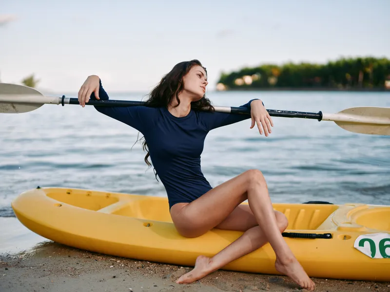 A young woman in a navy long-sleeve swimsuit sits on a bright yellow kayak on a sandy beach, stretching with a paddle across her shoulders under calm coastal light.