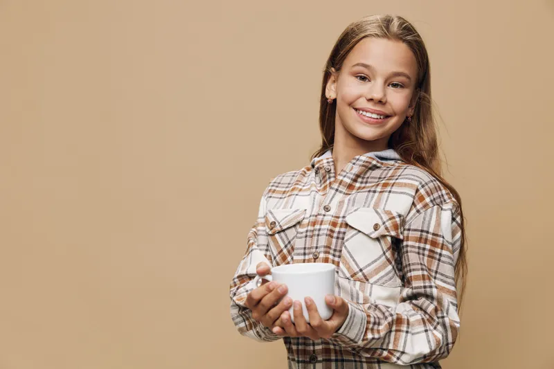 Girl smiling and holding white cup standing against beige background indoors. Child in plaid shirt enjoying hot beverage and looking at camera with cheerful expression.