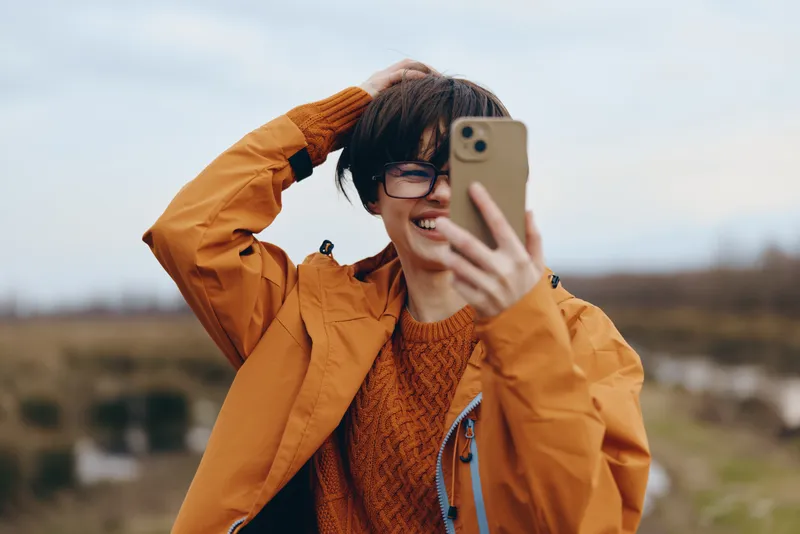 Woman outdoors taking selfie with smartphone, wearing orange jacket and glasses, smiling, autumn countryside background, natural daylight, casual portrait lifestyle, close view today