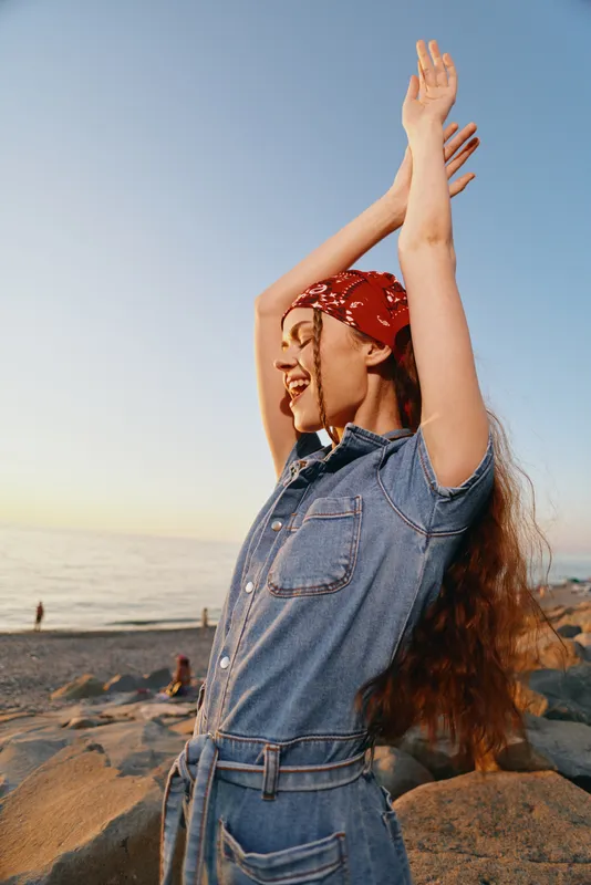 smiling woman in modern boho-western denim street style with long hair and red bandana, enjoying beach sunset with film color warm tones and relaxed lifestyle vibe