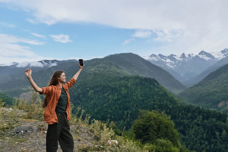 Woman hiker on a mountain ridge raises her arms and phone, capturing a vast landscape and outdoor panorama amid nature and distant peaks