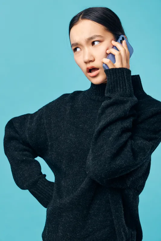 Young woman wearing black sweater talking on smartphone with annoyed expression isolated on blue background. Female person showing disbelief or confusion during phone call indoors.