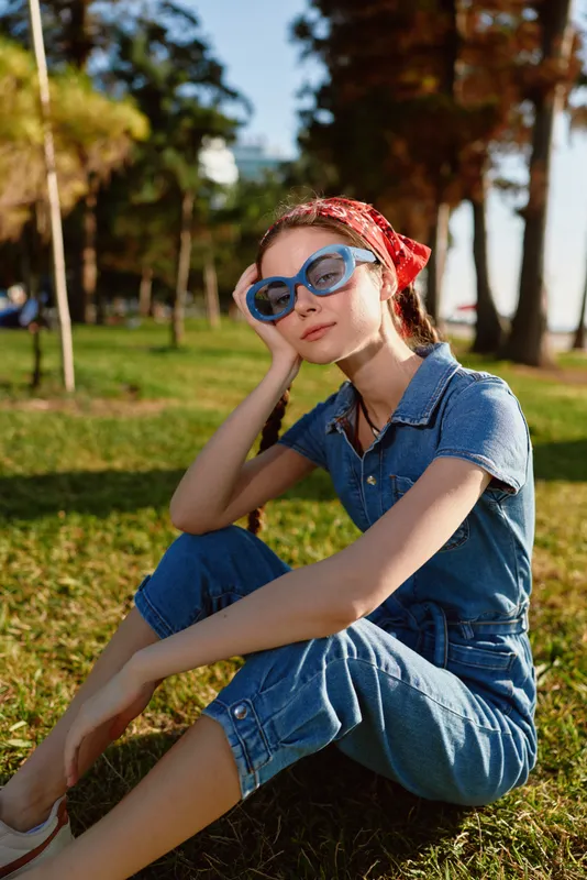 young woman wearing blue denim jumpsuit and sunglasses sitting on grass in sunny park with red headscarf and relaxed expression enjoying outdoors