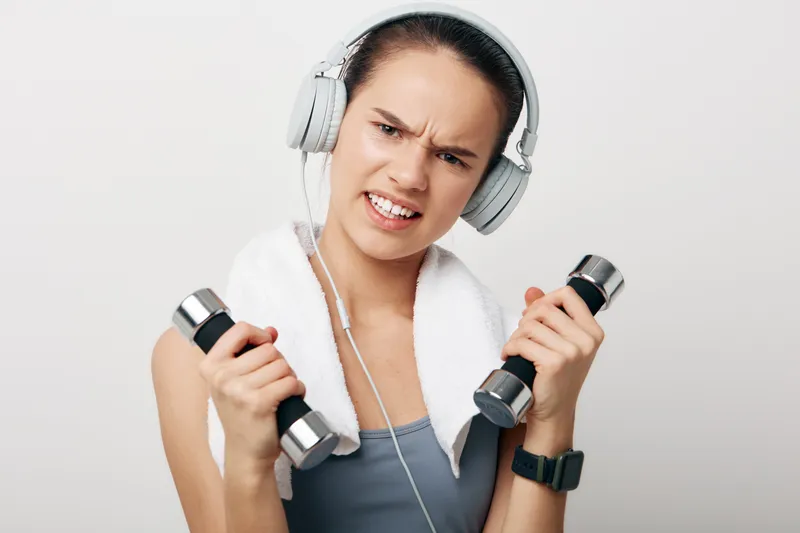 Focused woman in athletic apparel lifts dumbbells, a towel over her shoulders and large headphones, during a high-energy workout in a bright studio setting.
