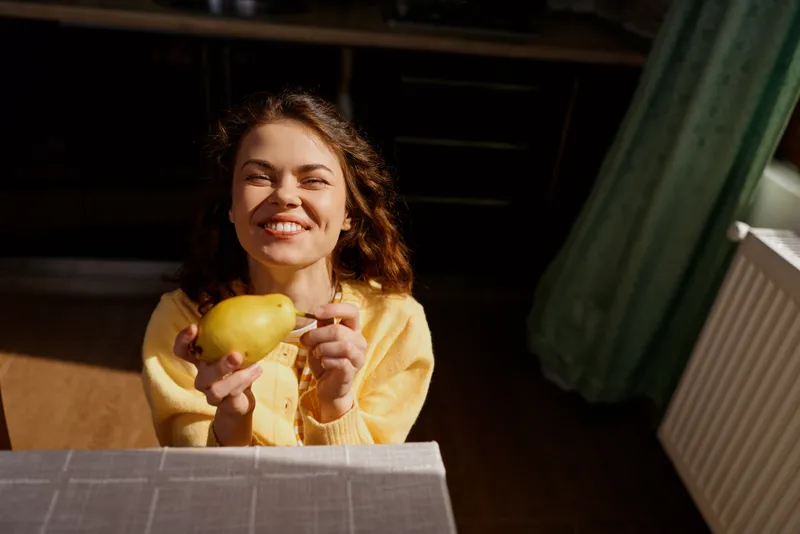 A smiling woman in a yellow sweater offers a mango at a cozy dining table, capturing a warm home lifestyle moment and casual happiness in a bright indoor space, inviting wellness and comfort.