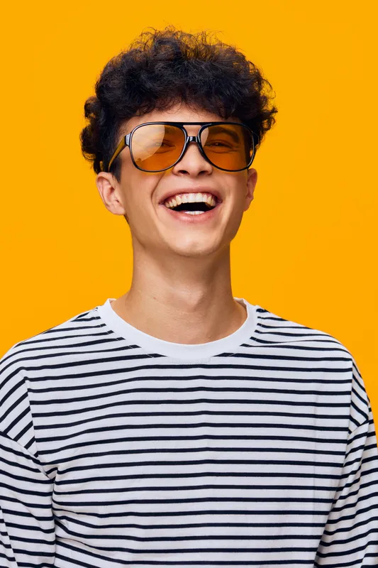 smiling young man wearing aviator sunglasses and a striped shirt, cheerful expression against bright orange background, casual style with curly hair