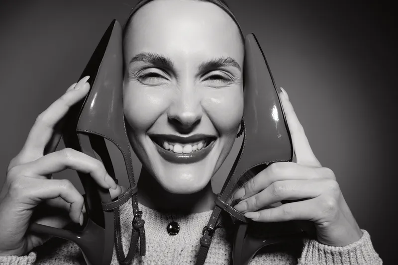 Smiling white woman with fair skin holds red high heels near her face. Close-up studio portrait in black and white. Concept for fashion, happiness, and shoe love.