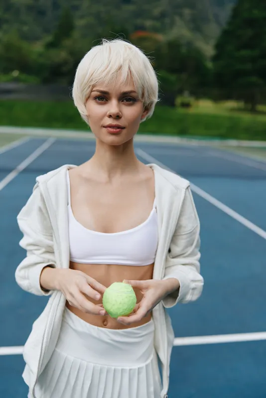 Young fit woman with short blonde hair in sportswear holding a tennis ball, exuding confidence on a sunny outdoor court in green surroundings
