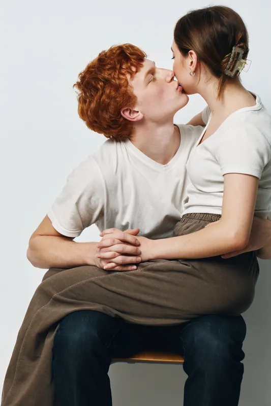 Couple in white shirts sitting on chair, young man holding girl on lap and kissing her cheek in intimate romantic moment with plain background.