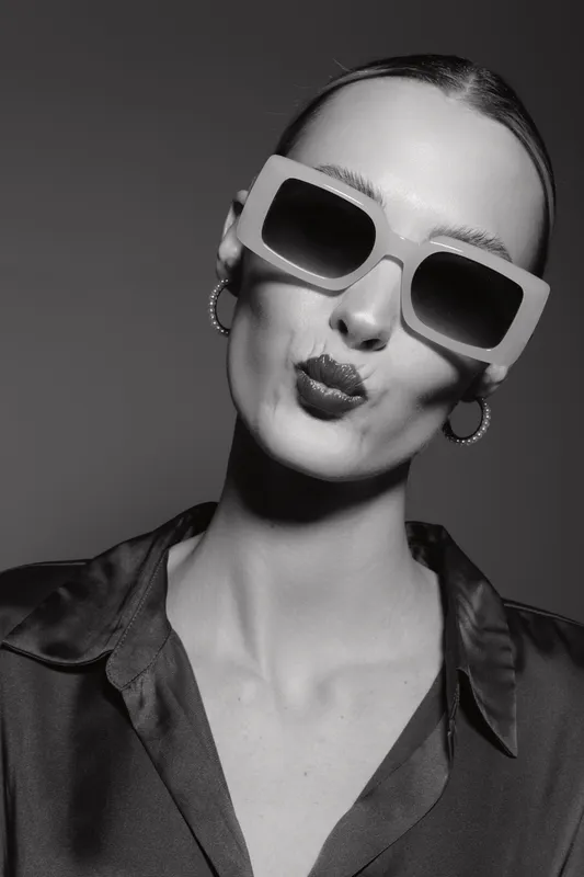 Slim white woman with pale skin wearing large sunglasses and hoop earrings makes a kiss face in studio.