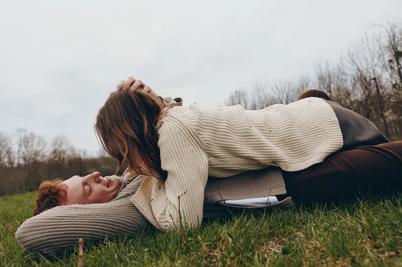 Couple outdoors, embracing on grass in cozy sweaters, candid moment of affection, sunlit air, relaxed posture, natural colors, soft breeze, serene countryside backdrop
