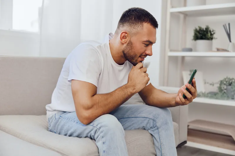 Thoughtful man engaging with smartphone, exploring digital world, comfortable living room setting with modern decor, showcasing contemporary lifestyle.