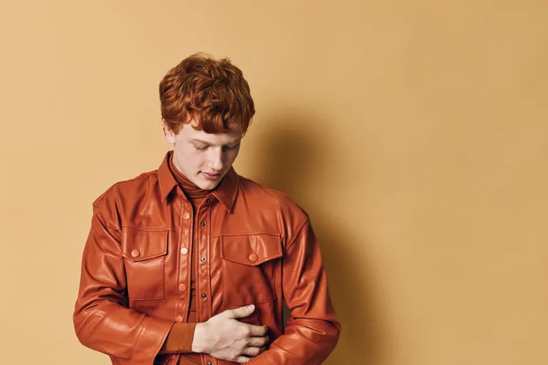 Young man with red hair wearing brown leather jacket standing against plain background looking down with thoughtful expression and folded arms.