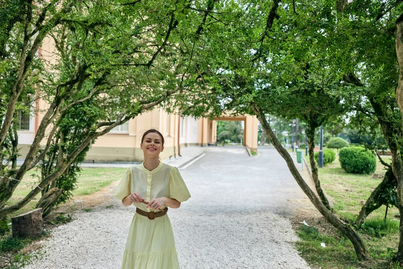 Woman smiling in light dress under park trees on gravel path outdoors authenticity inclusive portrait peaceful walkway scene