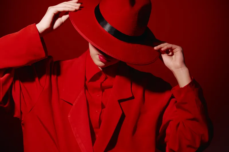 Fashion model wearing bright red outfit and stylish hat covers face with hands against vibrant red background. Concept of beauty, creative fashion, and artistic glamour.