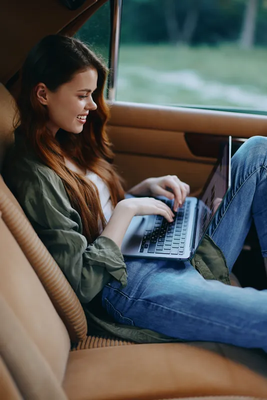 Young woman working on a laptop inside a vintage car, showcasing a relaxed mood, natural light, and a casual outfit of jeans and a top with a jacket.