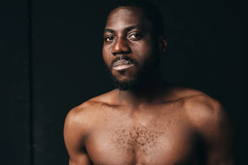 African man with dark skin and beard posing shirtless in creative conceptual visual style with serious expression and studio lighting on black background.