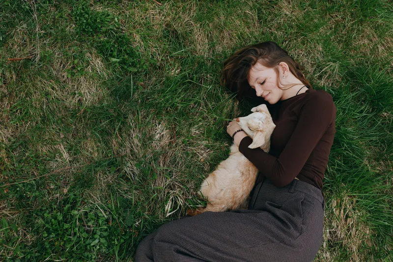 Relaxed woman lying on grass cuddling a goat kid, outdoor lifestyle moment, soft natural light, calm mood, connection and care, casual clothing, peaceful countryside vibe, intimate portrait