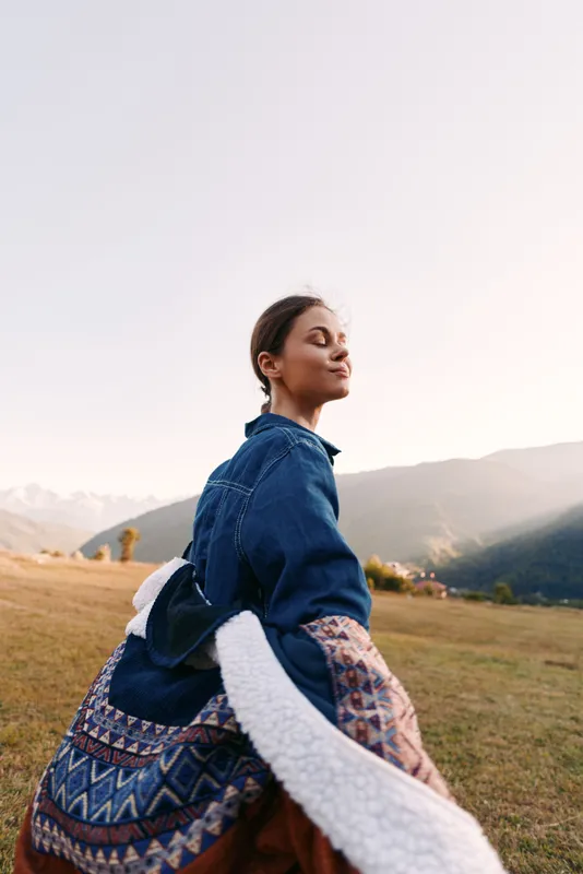 Woman portrait in nature with mountains and meadow, sunlight on her face as she breathes fresh air, eyes closed and relaxed wearing a denim jacket and patterned skirt at golden hour.