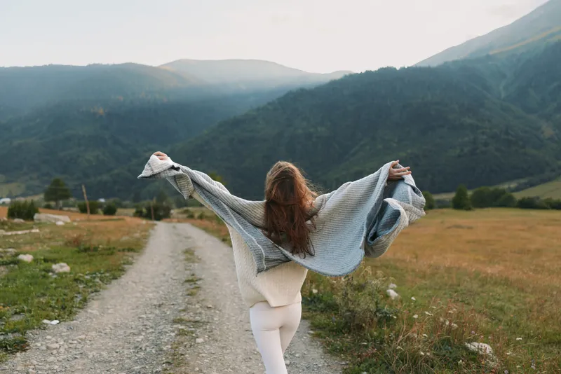 Young woman walks along a dirt path in a valley, arms wide with a scarf, expressing freedom and relaxation in a rustic landscape, her long hair flowing as distant mountains rise in warm morning light.