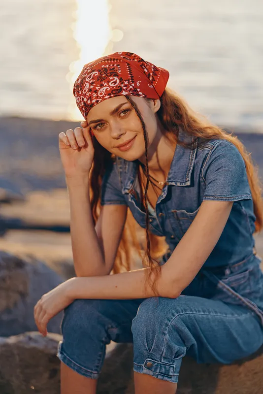 Lifestyle woman wearing modern boho-western denim street style with a red bandana, casual pose near water, film color tone creating warm, natural light atmosphere.