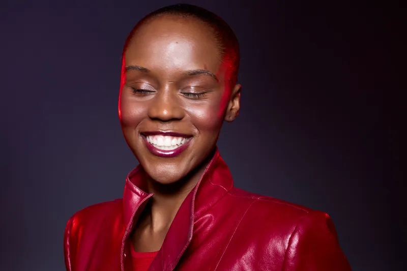 Smiling African woman with dark skin wearing a red leather jacket and lipstick poses with closed eyes on dark background. Concept of beauty, fashion, confidence, and elegance.