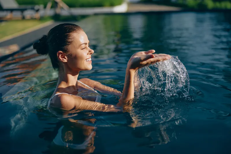 A smiling woman enjoys a playful splash in a bright pool, capturing a relaxed lifestyle moment outdoors under warm sunshine, refreshing water, leisure, wellness, and casual summer recreation in a