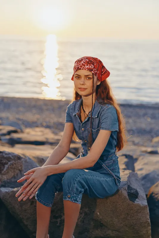 young woman sitting on rocks wearing denim outfit and red bandana by the sea during warm sunset light with calm ocean background and relaxed mood