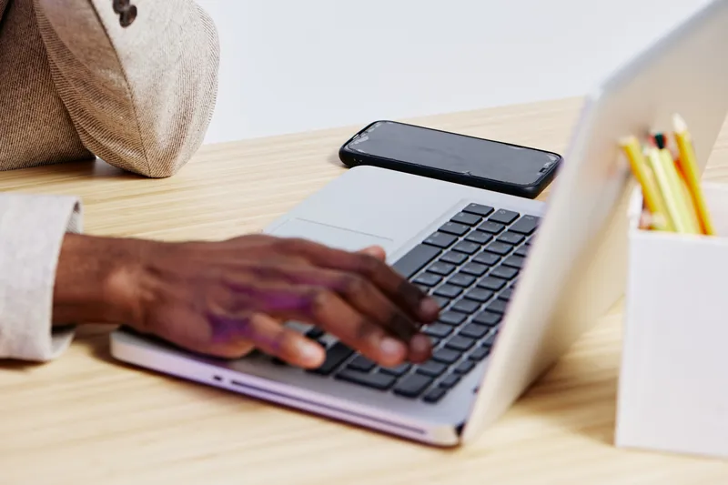 Close-up of a person working on a laptop with a smartphone and stationery on a wooden desk, representing modern office environment and remote work, lifestyle concept.